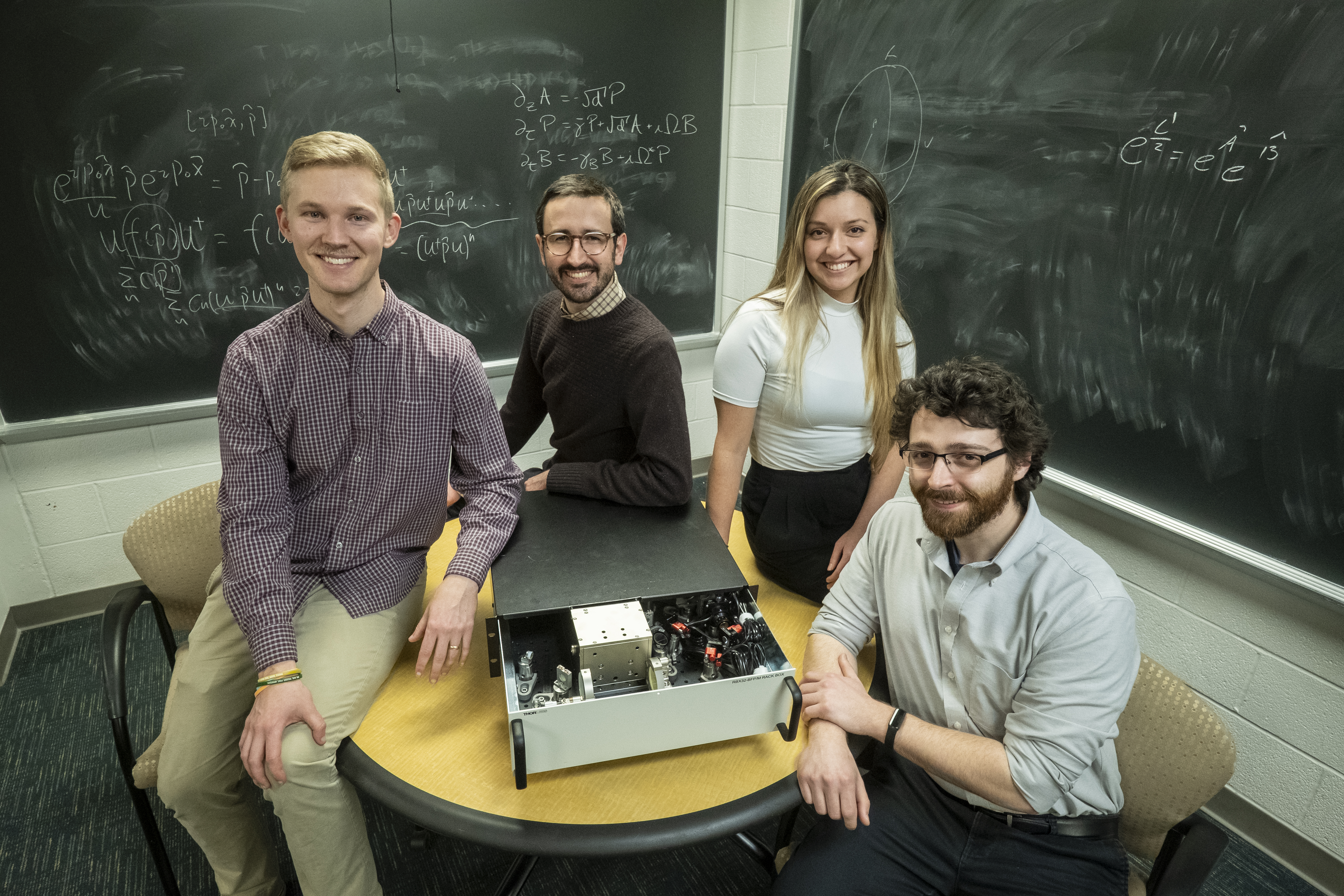 Three men and a woman stand in front of a chalkboard with equations on it, a computer device on the table between them.